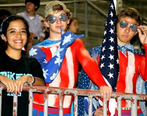 three students — a girl, and two boys who have painted themselves red, white, and blue — standing in the bleachers at a game
