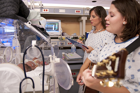 UA music therapy student Ellyn Hamm, holding guitar, sings to an infant while Cevasco charts.