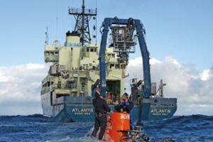Research Vessel Atlantis and the human-occupied vehicle Alvin are shown. Divers meet the sub when it surfaces to attach it to a hoist rope and bring it back on board (Image Woods Hole Oceanographic Institution).