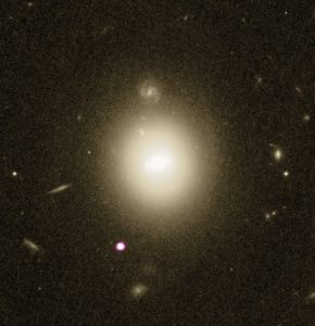 A NASA/ESA Hubble Space Telescope view of galaxy 6dFGS gJ215022.2-055059 – the large white-yellow blob at the center of the image – and neighboring galaxies, combined with X-ray observations of a black hole at the galaxy’s outskirts – the small white-purple dot to its lower left – obtained with NASA’s Chandra X-ray observatory.