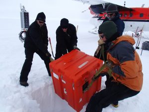 4 researchers lower an orange equipment box into a hole in the snow and ice